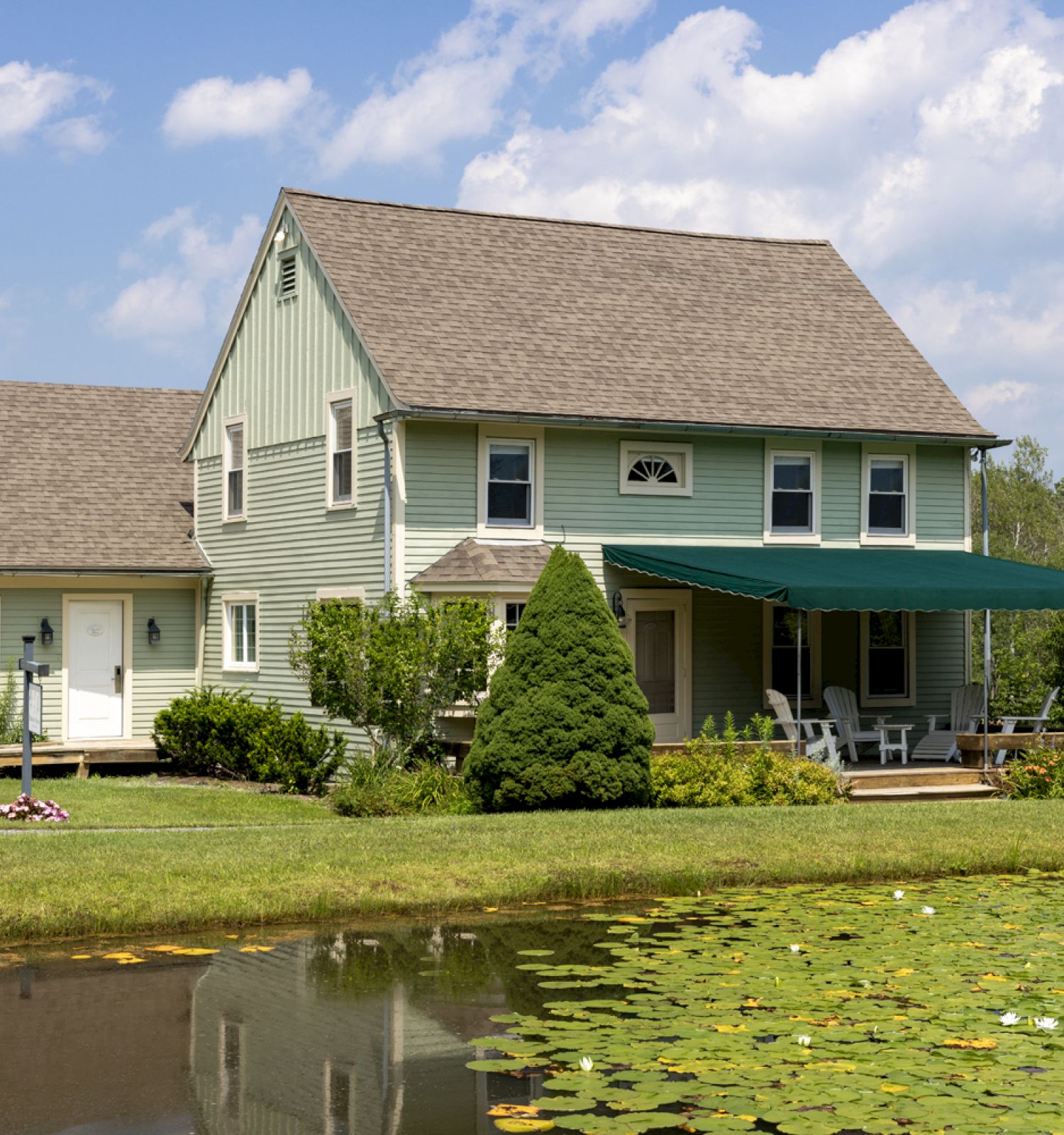 A two-story house with light green siding, a porch with green awning, and a pond with lily pads in the foreground is shown.
