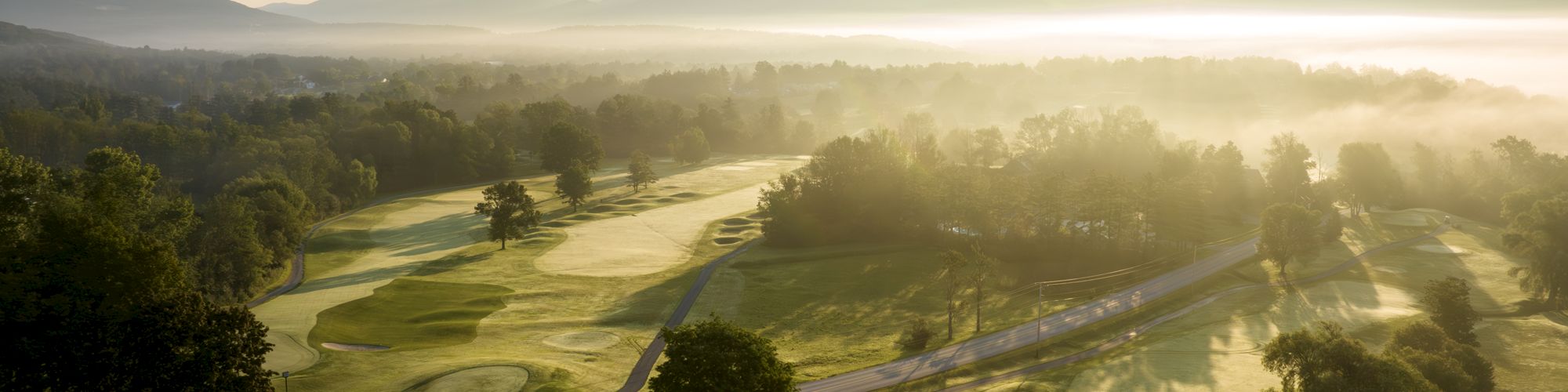 The image shows a serene golf course during sunrise with rolling hills in the background, a road, and some buildings in the foreground, bathed in morning light.