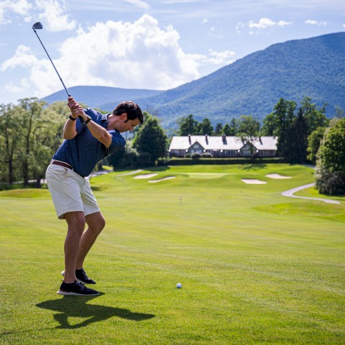 A person playing golf on a lush green course with mountains and a clear blue sky in the background.