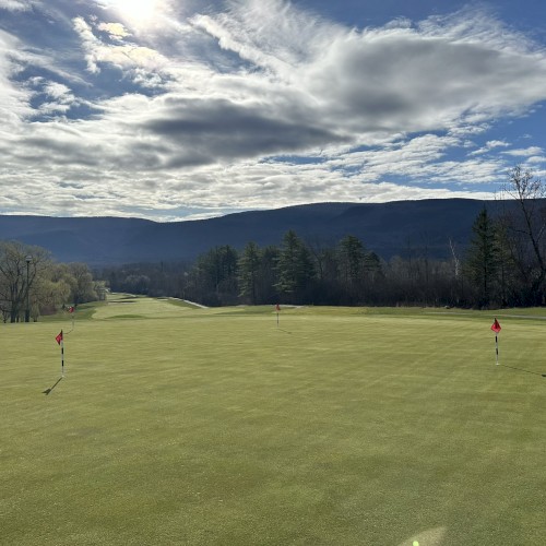 The image shows a golf course with multiple flags on the green, surrounded by trees and hills under a partly cloudy sky.