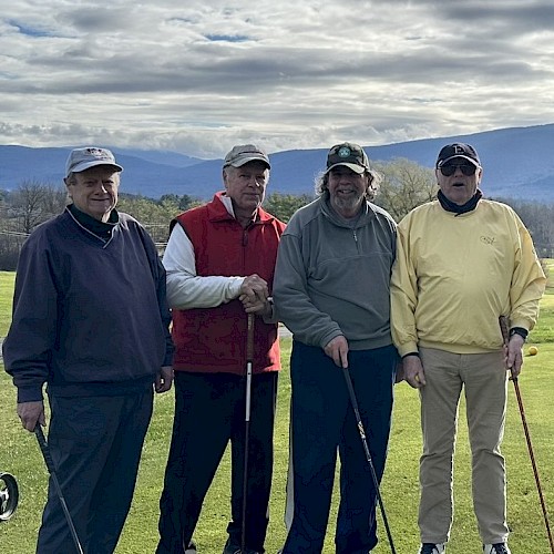 Four men are standing on a golf course with golf clubs, mountains and a cloudy sky in the background, all wearing different colored outfits and hats.