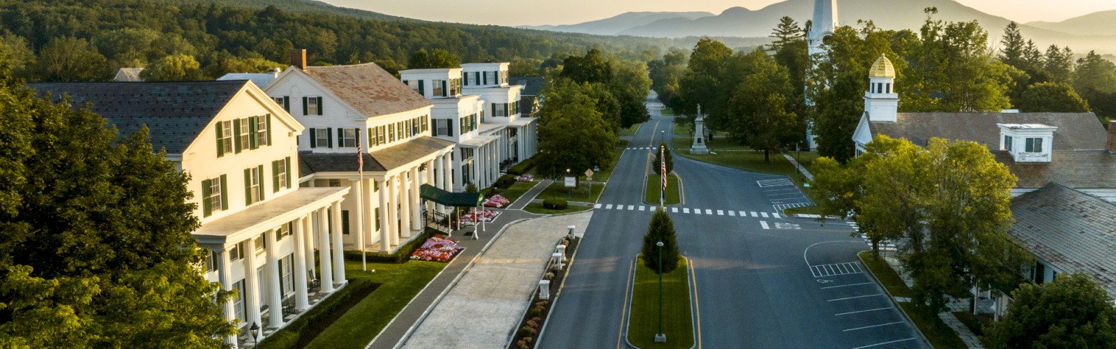 An aerial view of a quaint small town with classic white buildings, a church with a steeple, tree-lined streets, and mountains in the background.