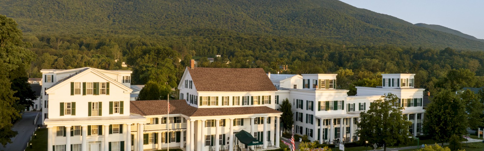 A large, white building complex surrounded by trees and mountains in a serene landscape, with a clear blue sky in the background.