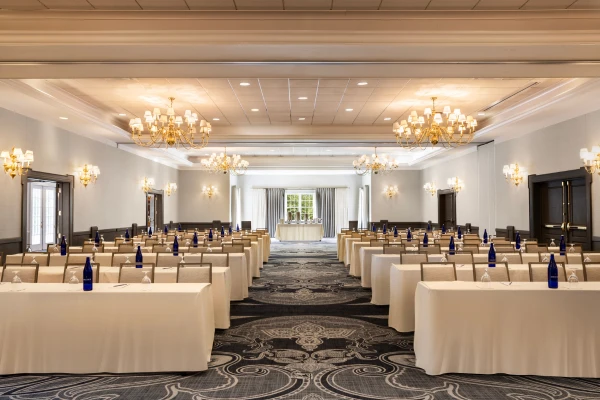 This image shows an elegant conference room set up with rows of tables and chairs, each with blue water bottles. Chandeliers hang from the ceiling.