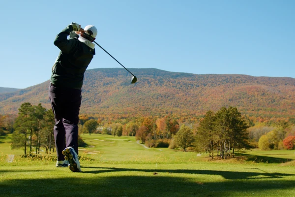 A person is swinging a golf club on a scenic golf course with vibrant fall foliage and mountainous background under a clear blue sky.
