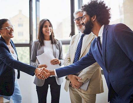 Four people in business attire are in an office setting, two shaking hands, all smiling and looking happy, with documents in the background.