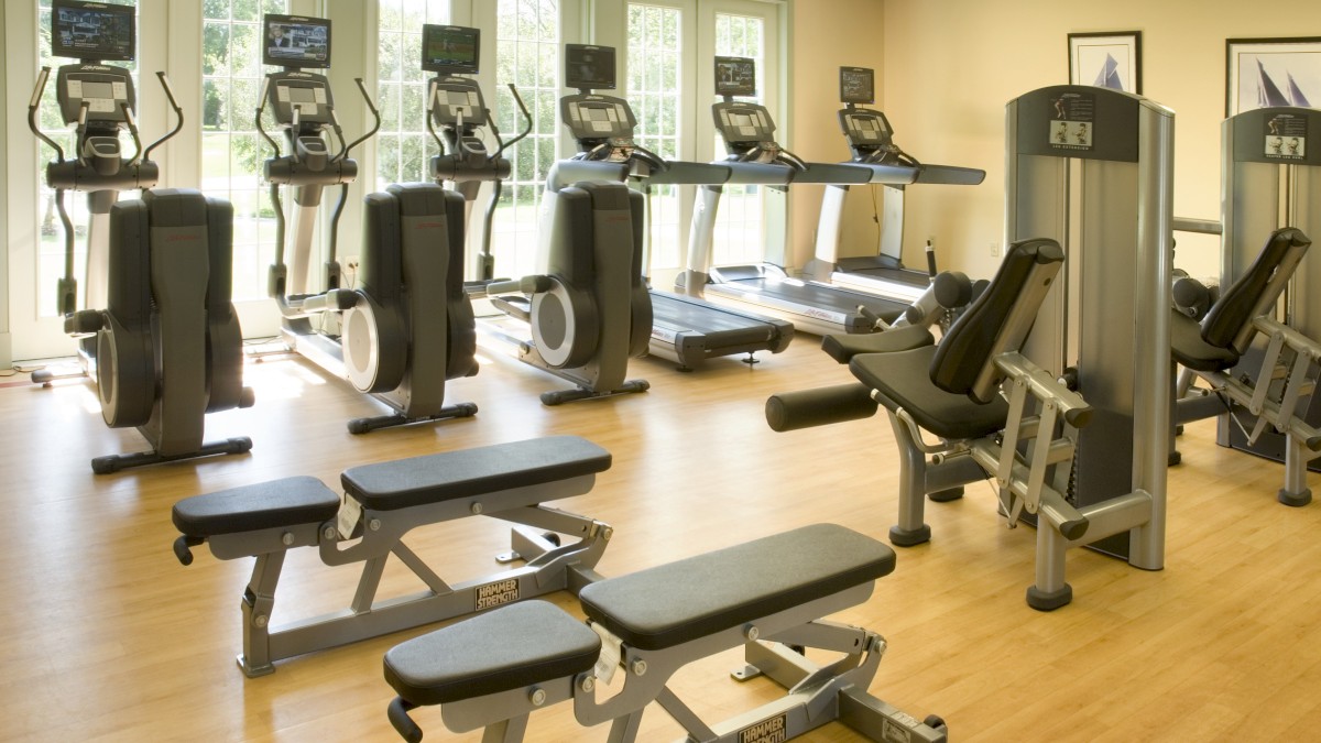 The image displays a well-equipped gym with exercise machines including treadmills, ellipticals, and weight benches, all set in a clean, spacious area.