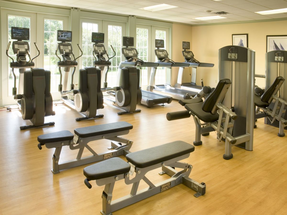 The image displays a well-equipped gym with exercise machines including treadmills, ellipticals, and weight benches, all set in a clean, spacious area.