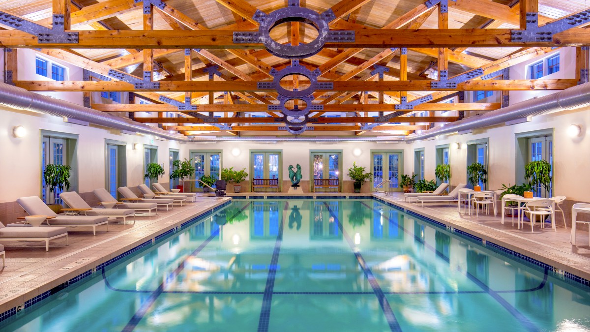 This image shows an indoor swimming pool area with wooden beams on the ceiling, lounge chairs, potted plants, and tables with chairs on the side.