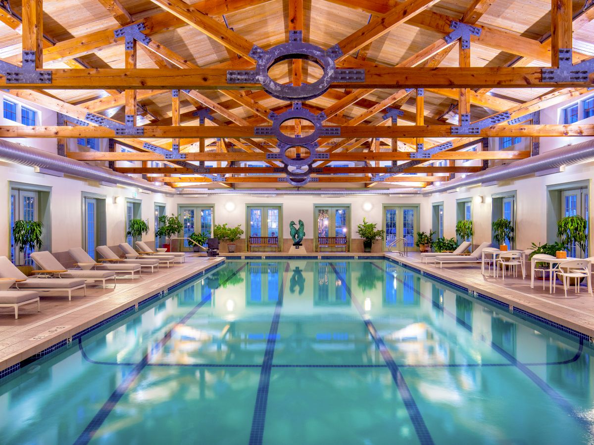 This image shows an indoor swimming pool area with wooden beams on the ceiling, lounge chairs, potted plants, and tables with chairs on the side.