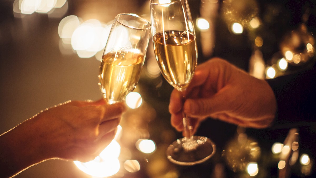 Two hands holding champagne flutes, clinking together in a celebratory toast with a blurred, festive background of lights.