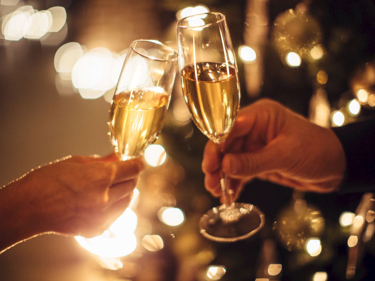 Two hands holding champagne flutes, clinking together in a celebratory toast with a blurred, festive background of lights.