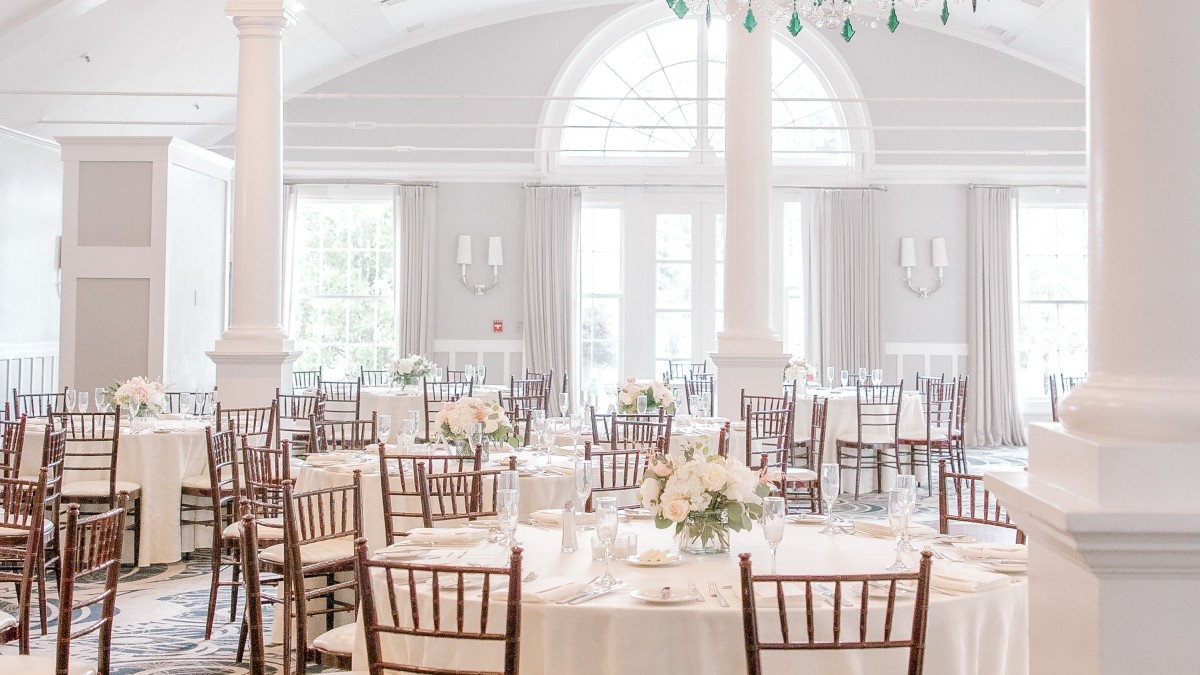 An elegant event venue with round tables, wooden chairs, white tablecloths, floral centerpieces, chandeliers, and large windows with arched tops.