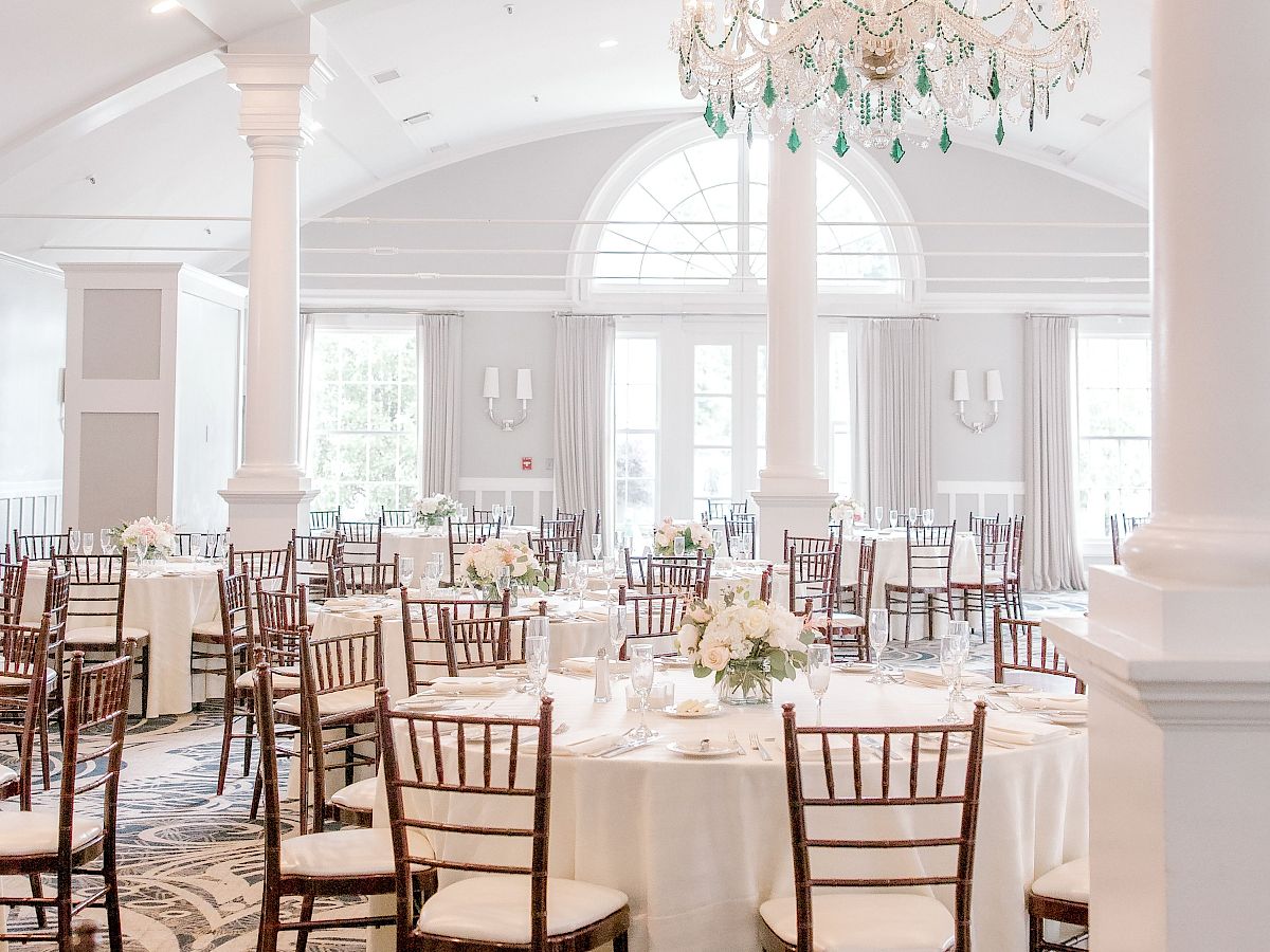 An elegant event venue with round tables, wooden chairs, white tablecloths, floral centerpieces, chandeliers, and large windows with arched tops.