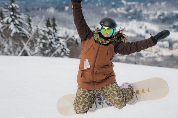 A person in winter gear is kneeling on a snowy slope with arms raised, holding a snowboard, and mountains and trees in the background.