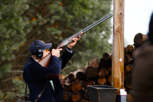 A person is aiming a shotgun upwards outdoors, equipped with ear protection and a cap, standing by a pile of wood logs.