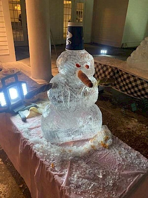 The image shows an ice sculpture of a snowman with a carrot nose, red eyes, and a black hat on a pink cloth-covered table at night.