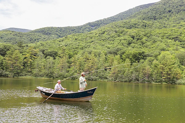 Two people are fly fishing from a small boat on a calm lake surrounded by lush, green, forested mountains under a cloudy sky.