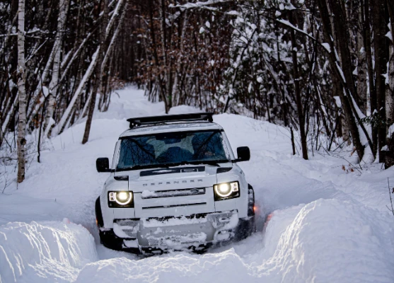 A white SUV navigates through a snowy forest trail with its headlights on.