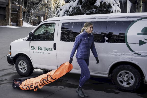 A person in winter clothing stands near a Ski Butlers service van, pulling a large orange ski equipment bag on a snowy street.