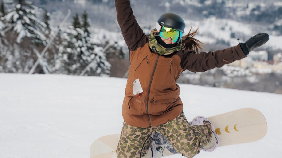 A person wearing winter gear and goggles kneels in snow with arms raised in excitement, holding a snowboard on a snowy mountain backdrop.