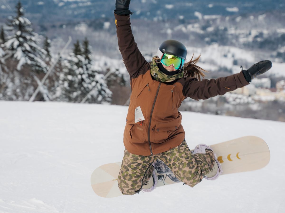 A person wearing winter gear and goggles kneels in snow with arms raised in excitement, holding a snowboard on a snowy mountain backdrop.