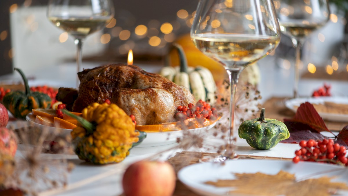 A festive table setting with a roast, wine glasses, and decorative gourds and leaves, lit by fairy lights in the background.