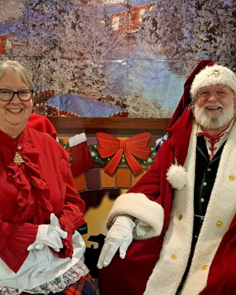 An elderly couple in festive attire sits in front of a winter-themed backdrop, featuring a snowy village.