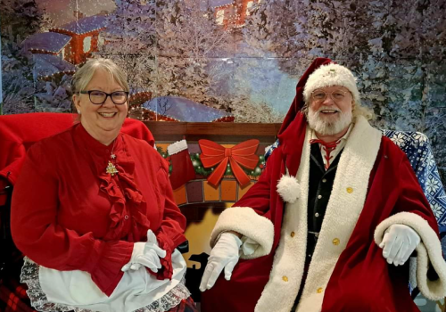 An elderly couple in festive attire sits in front of a winter-themed backdrop, featuring a snowy village.