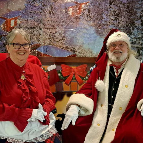 An elderly couple in festive attire sits in front of a winter-themed backdrop, featuring a snowy village.