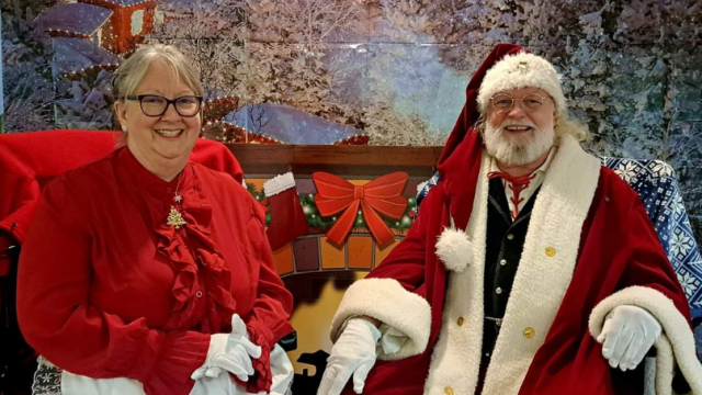 An elderly couple in festive attire sits in front of a winter-themed backdrop, featuring a snowy village.