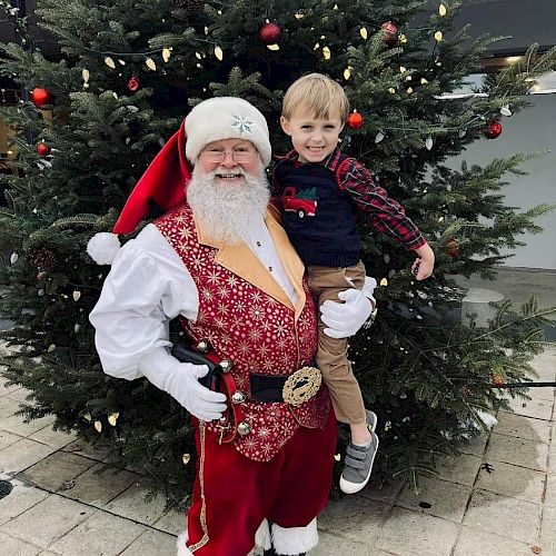 A person dressed as Santa Claus is holding a smiling child in front of a decorated Christmas tree.