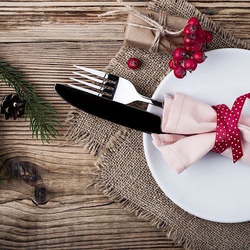 A festive table setting with a white plate, a fork, and a knife wrapped in a pink napkin tied with a red ribbon, alongside pine leaves and berries.