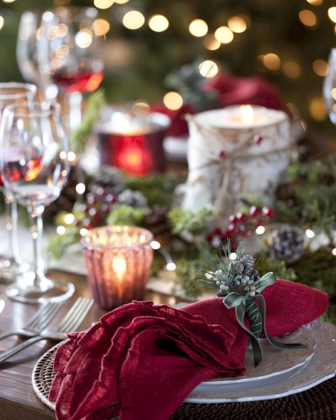 A festive dinner table setup with red napkins, wine glasses, candles, and holiday decorations, including greenery and lights.