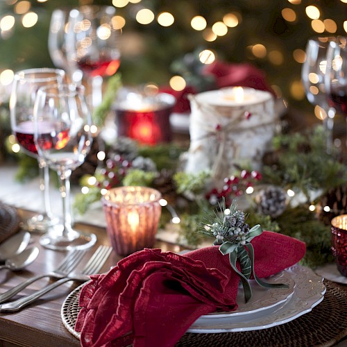 A festive dinner table setup with red napkins, wine glasses, candles, and holiday decorations, including greenery and lights.