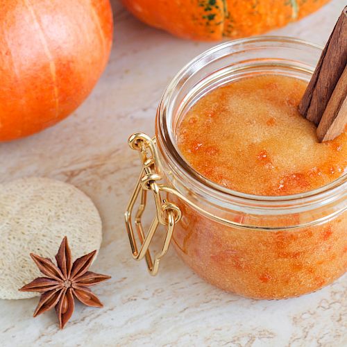 A jar filled with an orange substance, possibly pumpkin-based, with cinnamon sticks. Nearby are whole pumpkins, a star anise, and a round stone.