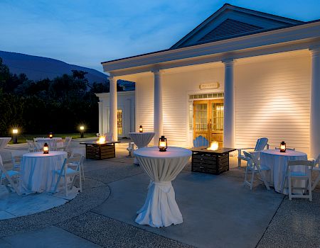Outdoor evening event setup with high tables and lantern lights in front of a white building with columns.