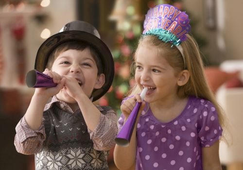 Two children are celebrating, wearing party hats and blowing noisemakers. There are festive decorations in the background ending the sentence.