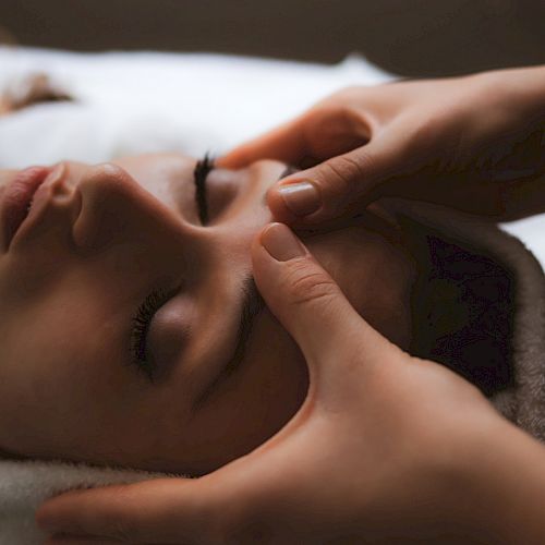 A person is receiving a relaxing face massage, with their eyes closed and head resting on a towel while two hands gently press on their forehead.