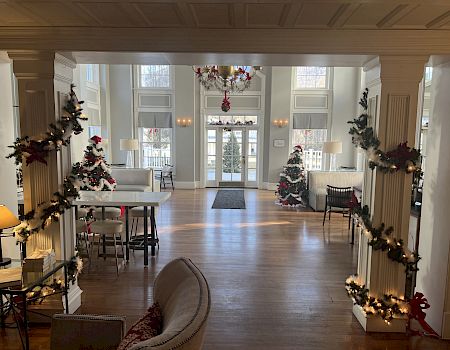 A festive, well-lit room with Christmas trees, garlands, and decorations, featuring a large entrance with windows, cozy seating, and hardwood floors.