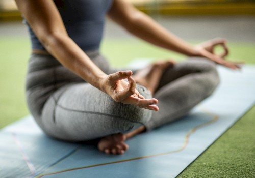 A person practices yoga in a cross-legged position on a mat, hand in a mudra, wearing athletic clothing, on a grass-like surface.