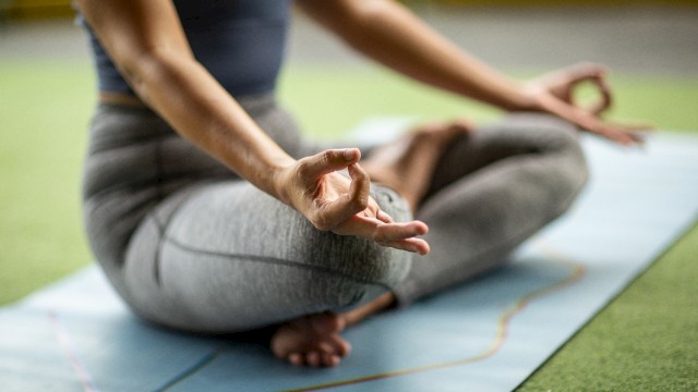 A person practices yoga in a cross-legged position on a mat, hand in a mudra, wearing athletic clothing, on a grass-like surface.