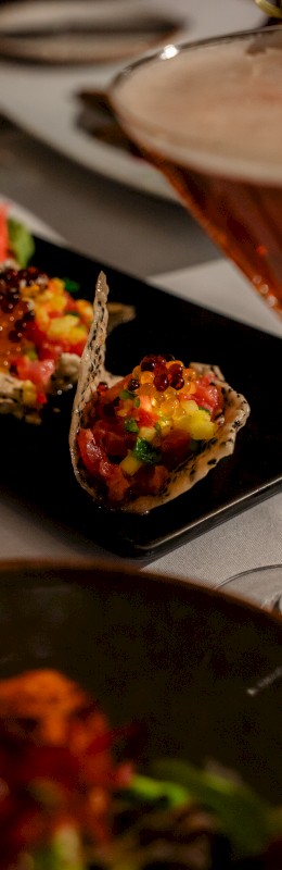 The image shows a dish with colorful appetizers, possibly served in crispy shells, next to a cocktail in a sleek glass on a dining table.