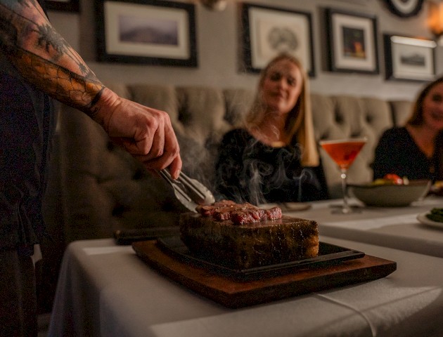 A person is serving a steak at a restaurant table. Two women are seated with cocktails and sides, and framed art decorates the wall.
