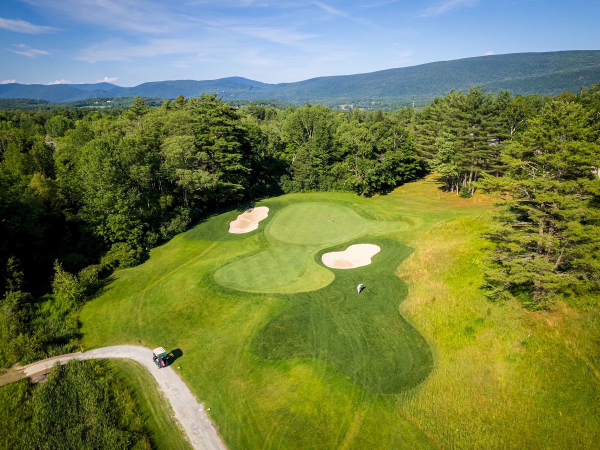 An aerial view of a golf course with sand bunkers, a green, a player, and a golf cart, surrounded by lush trees and distant mountains ending the sentence.