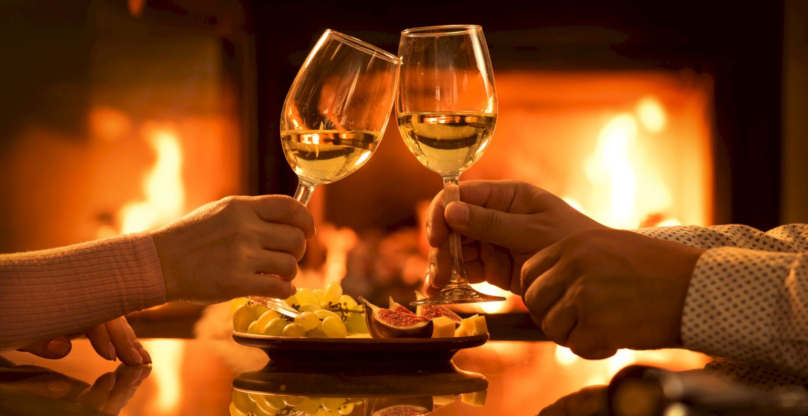 Two people clink glasses of white wine by a fireplace, with a plate of food in the foreground.