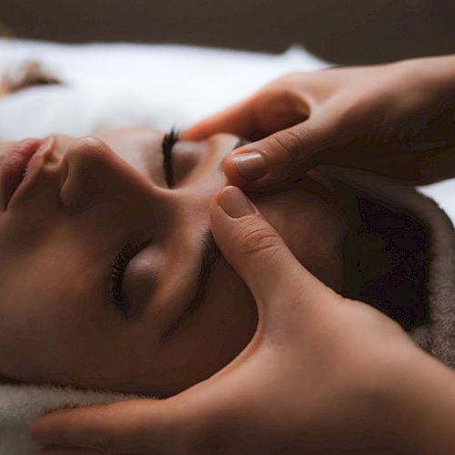 A person is receiving a facial massage, lying down with eyes closed, while wrapped in a towel, creating a calm and relaxing atmosphere.