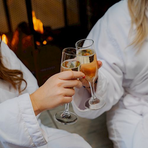 Two people in white robes clink champagne glasses by a cozy fireplace, celebrating a relaxing moment together.