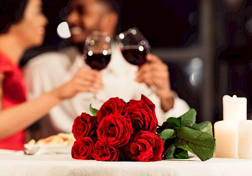 A couple toasts with glasses of red wine at a romantic candlelit dinner, with a bouquet of red roses on the table.