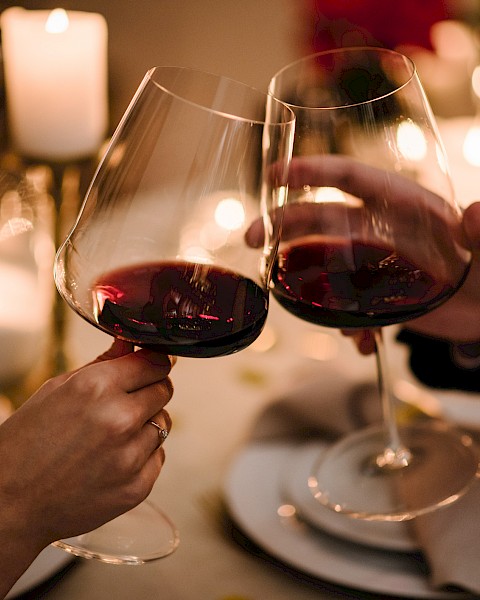 Two people clinking glasses of red wine at a candlelit table, surrounded by cozy, warm lighting and an elegant dinner setting.